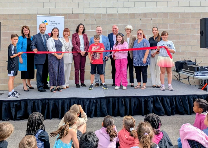 Des dignitaires, des membres du Conseil, la direction de l’école et des élèves procèdent à la coupure du ruban lors de l’inauguration officielle de l’École élémentaire catholique Marie-Soleil, sous le regard des élèves rassemblés devant la scène.